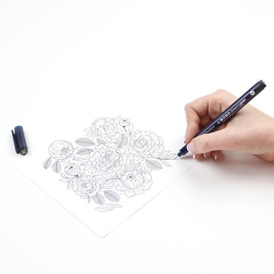 A hand holding a black Tombow Mono Black drawing pen, sketching a floral line art design on a white sheet of paper. The black cap of the pen lies nearby on the white surface.