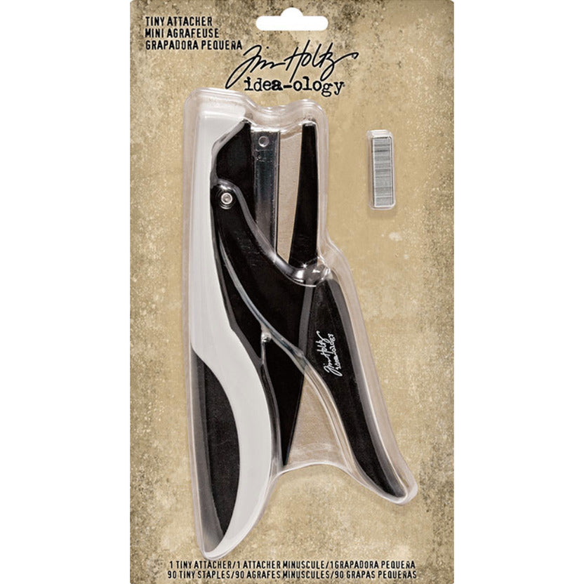 A Tim Holtz Mini Asbaise (Tiny Attacher) paper clip cutter in black and white, packaged in a clear plastic sleeve on a textured beige cardboard backing card.